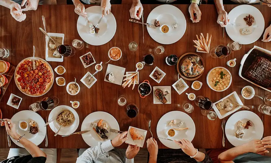 Une grande table partagée vue du dessus, plats et mains des convives